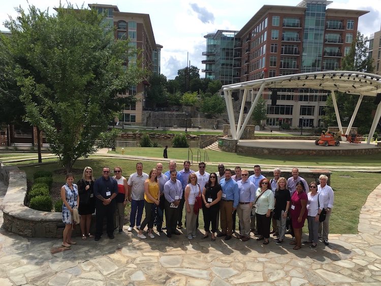 Fort Wayne business and civic leaders stop for a photo near a riverfront amphitheater and RiverPlace, a development that includes apartments, retail and office space, and a Hampton Inn.