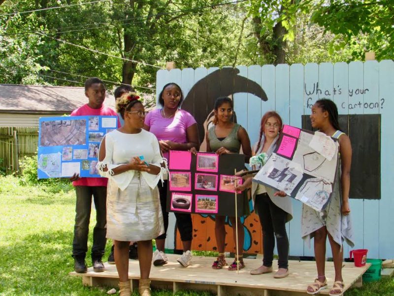 A Tired-a-Lot Summer Studio group presents their designs for a community space in front of a mural they designed and painted.