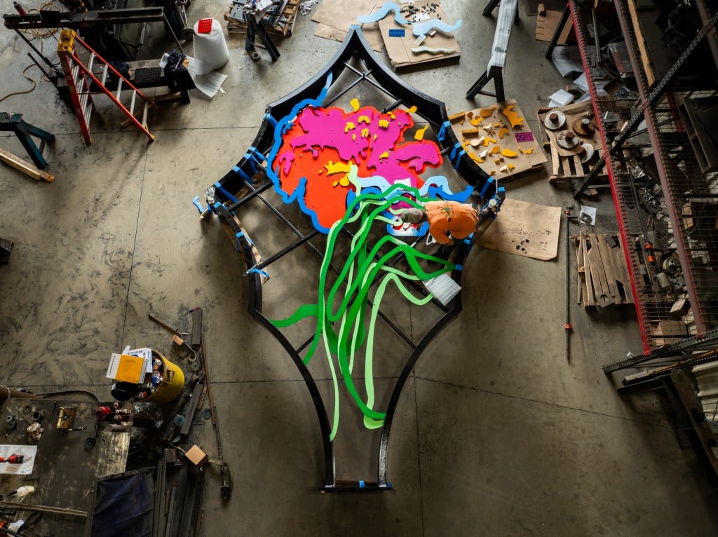 A large metal sculpture that is laying on the ground of a workshop. The sculpture is black, green, blue, pink, orange and yellow.