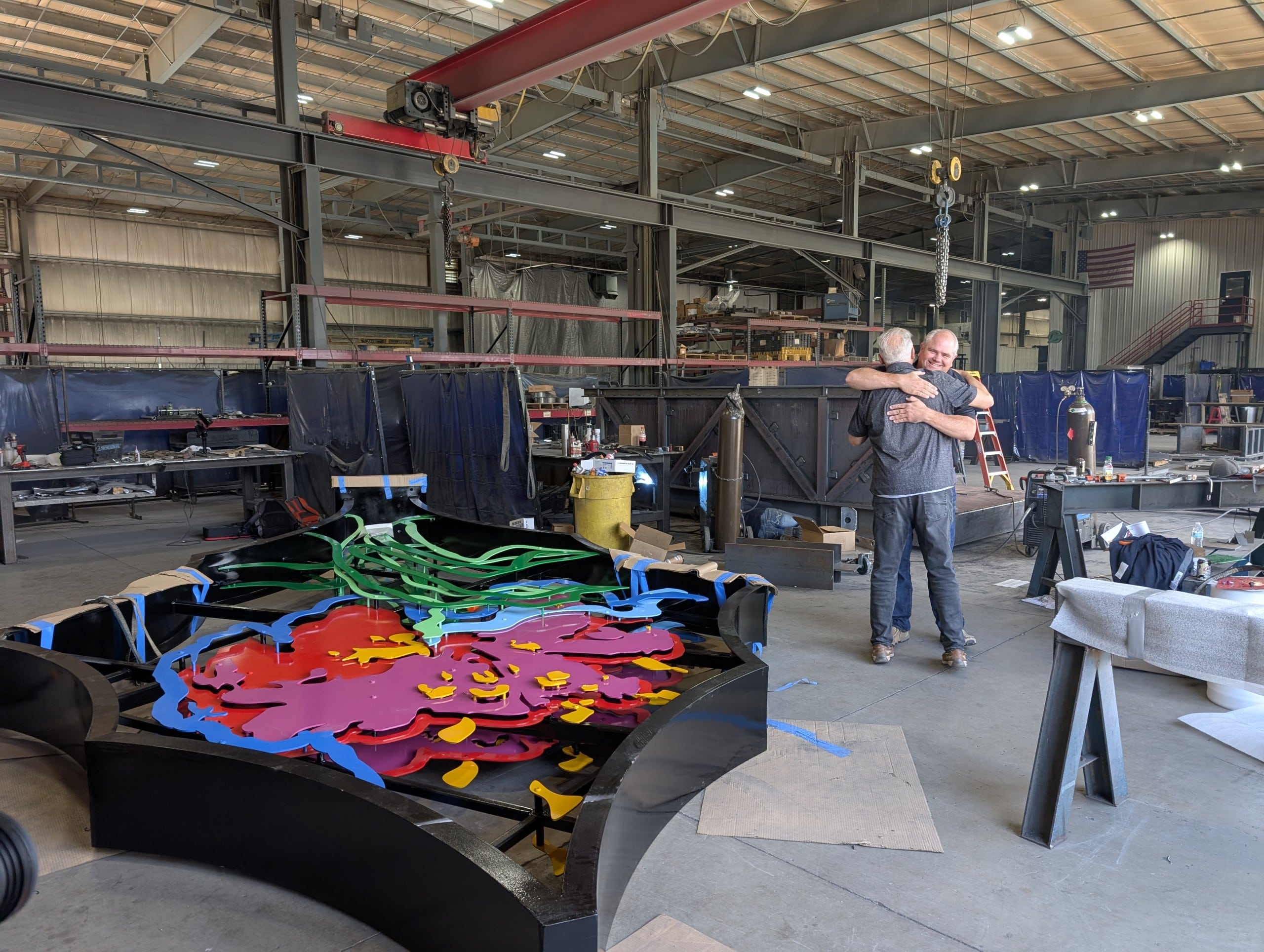 Inside a workshop, two men hug next to a large metal sculpture that is laying on the ground. The sculpture is black, green, blue, pink, orange and yellow.