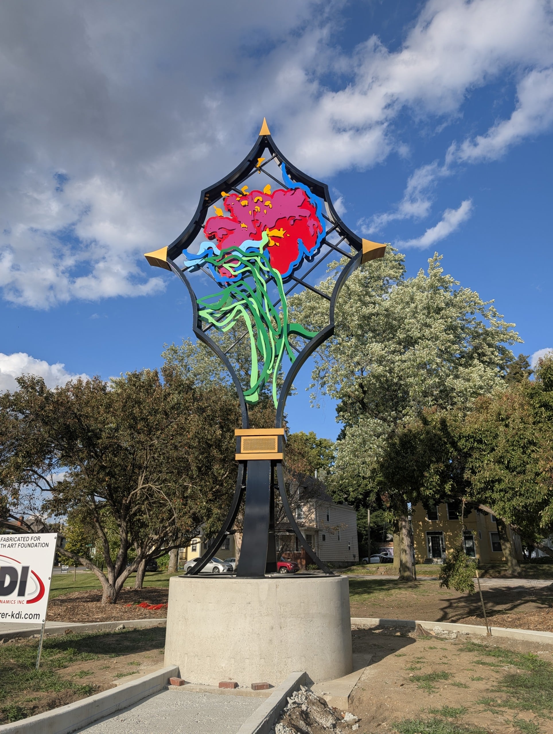 A large metal sculpture stands atop a circular concrete base. The sculpture is black, green, blue, pink, orange and yellow.