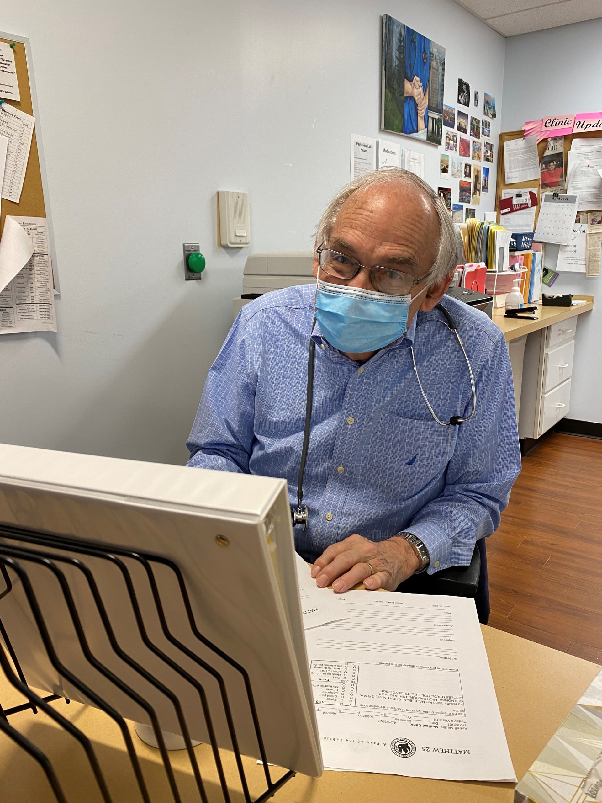 A male doctor in a blue button up shirt fills out paperwork at a desk.