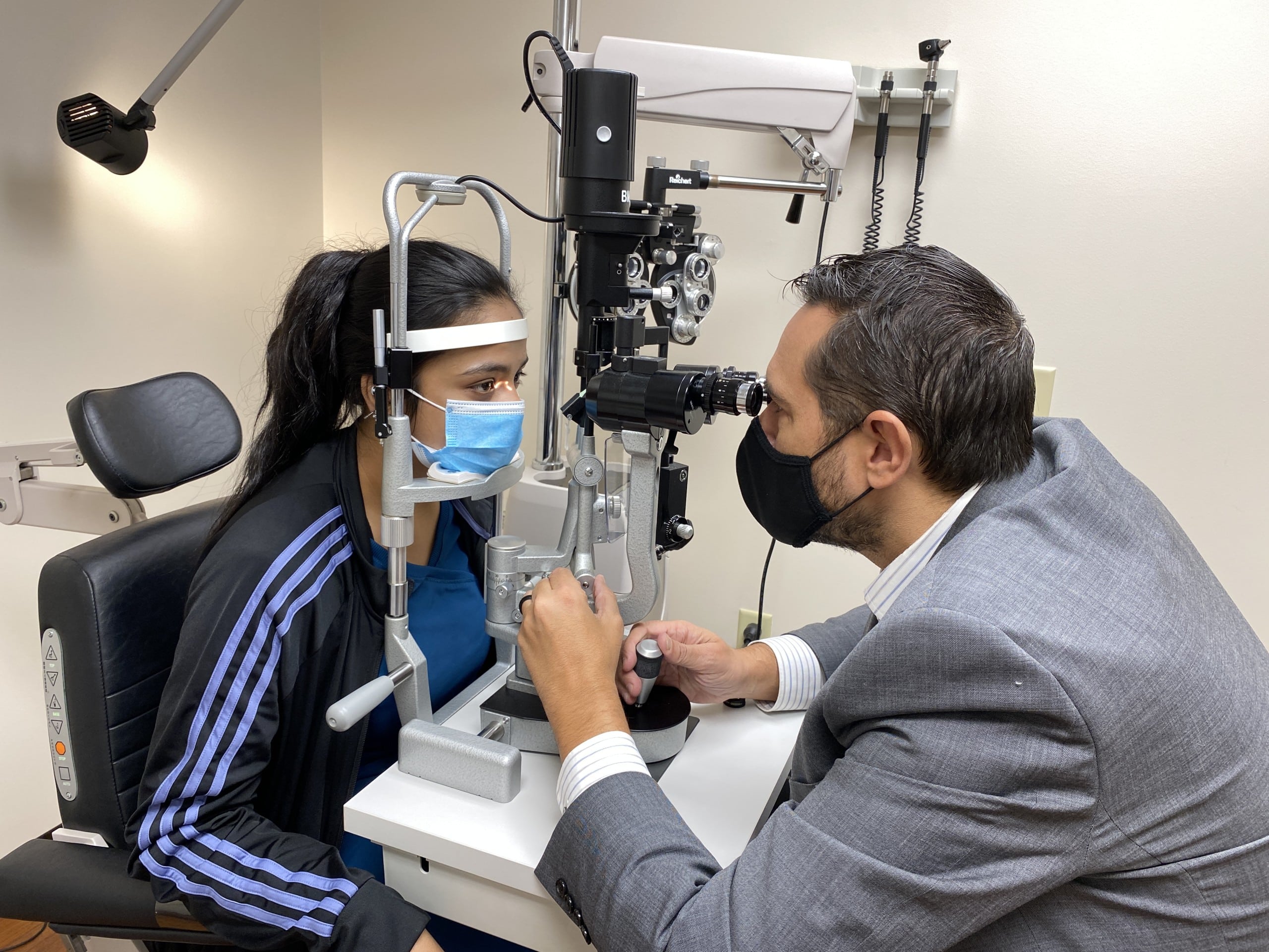 An eye doctor wearing a grey jacket looks at the eyes of a patient.