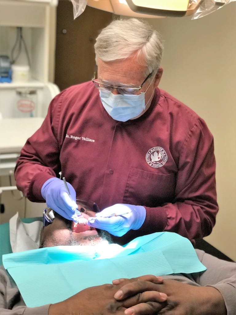 A dentist with tools in both hands examines the mouth of a patient who is laying on their back in an exam chair.