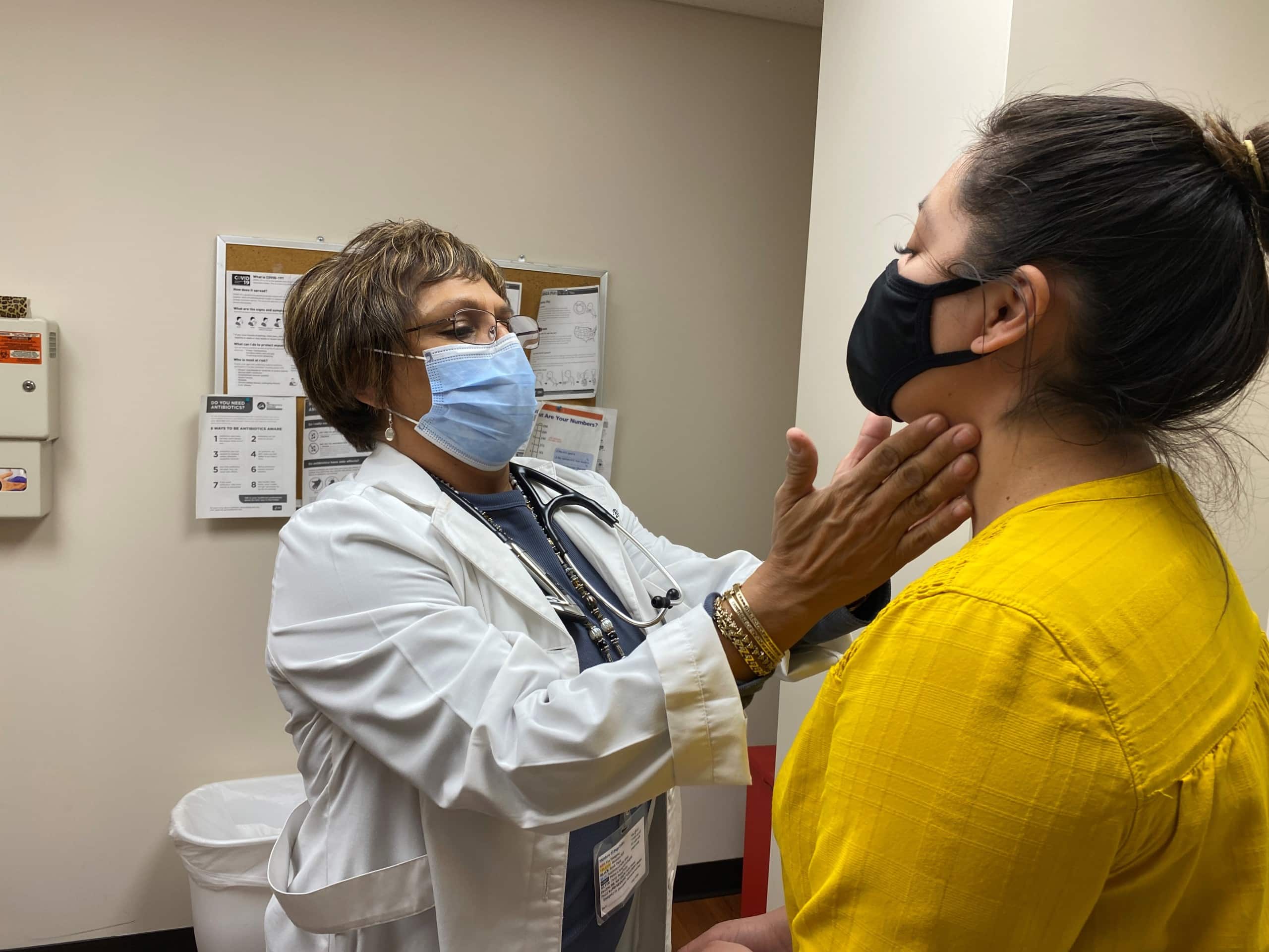 A doctor in a white coat and face mask touches the neck of a patient in an exam room.