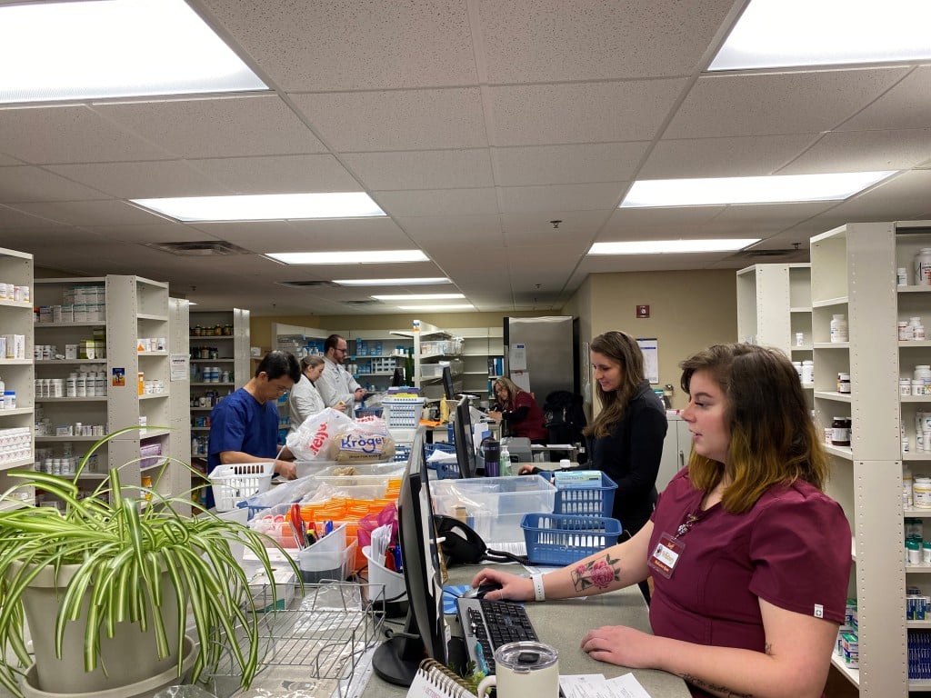 Six staff members stand at a long desk inside a pharmacy room.