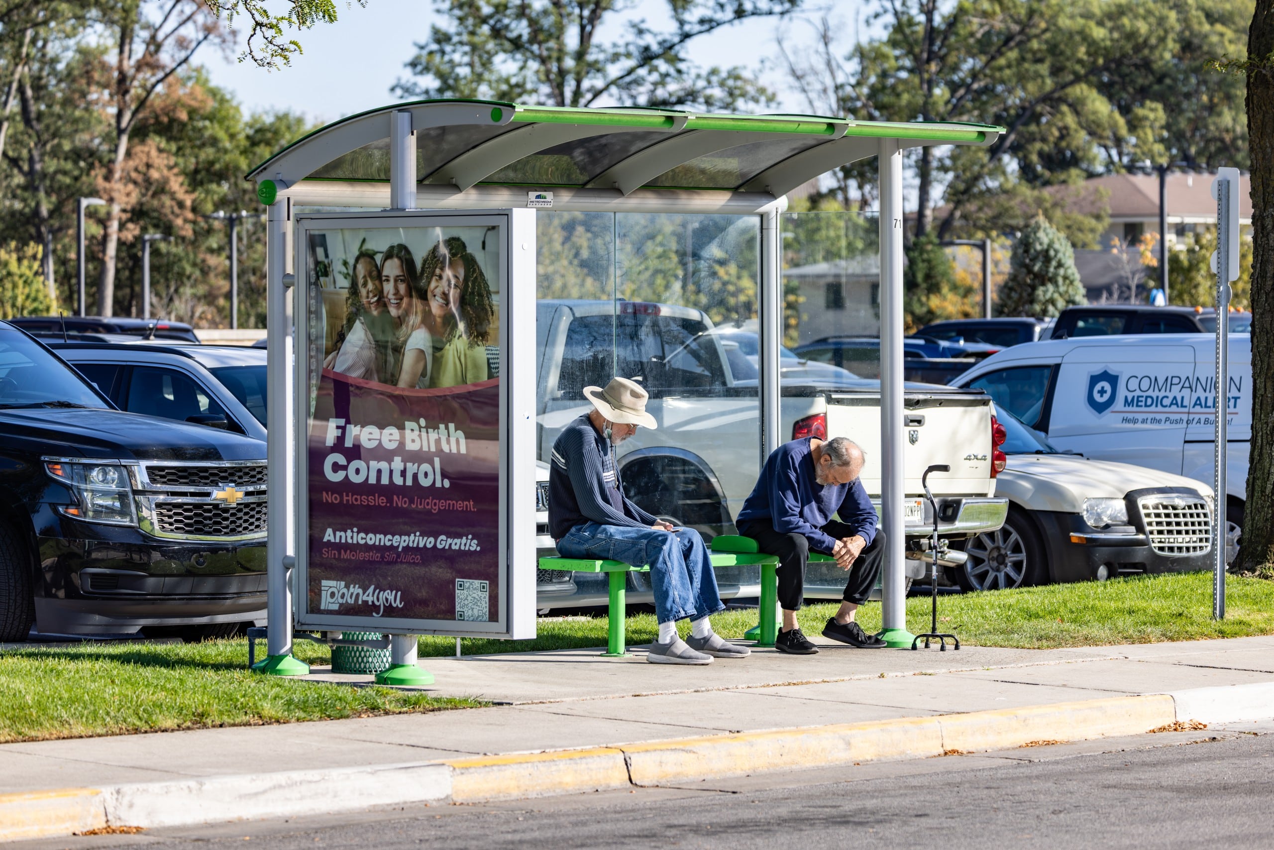 This shows a bus shelter with three glass walls. Inside two people sit and wait.