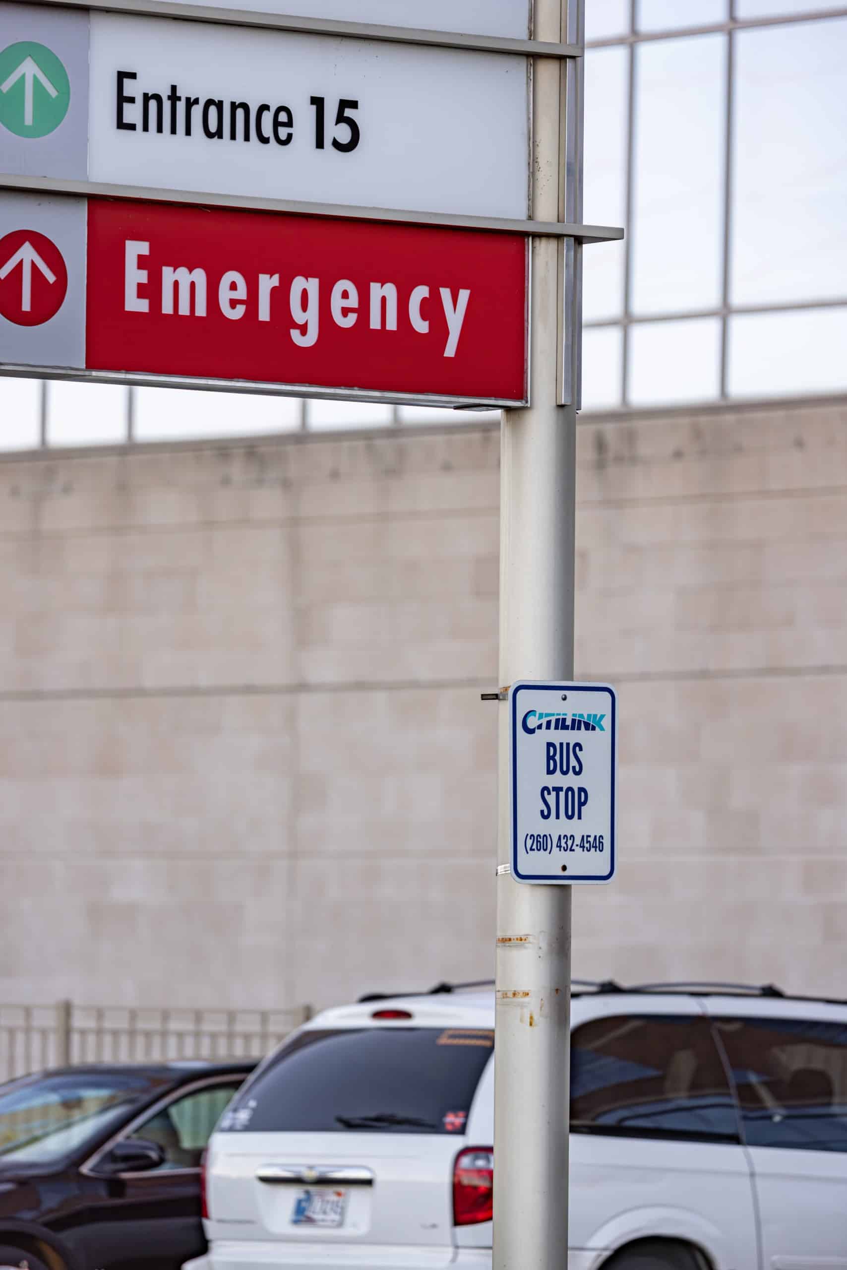 A round metal pole hosts two signs. A red sign reads, "Emergency" with an arrow pointing forward. A second white and blue sign reads, "Citilink Bus Stop".