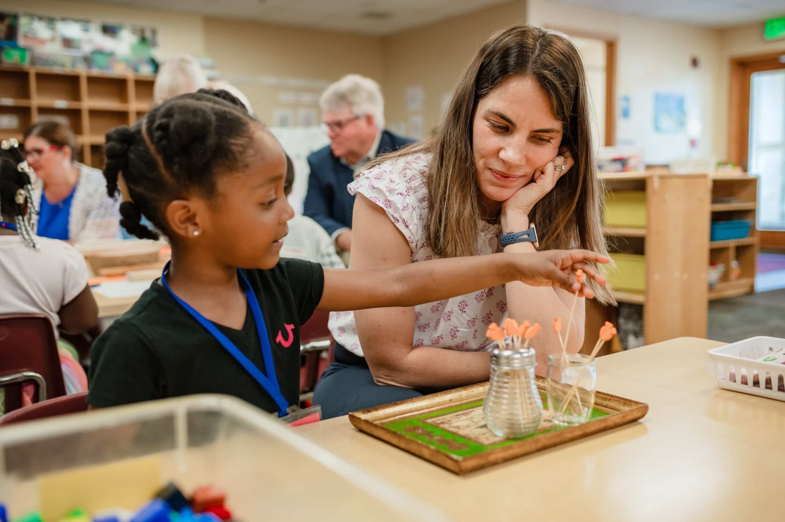 Inside a classroom for young students, a woman with long brown hair watches a young student, who is moving sticks from one to to another to learn counting. Both are sitting at a short wooden desk.