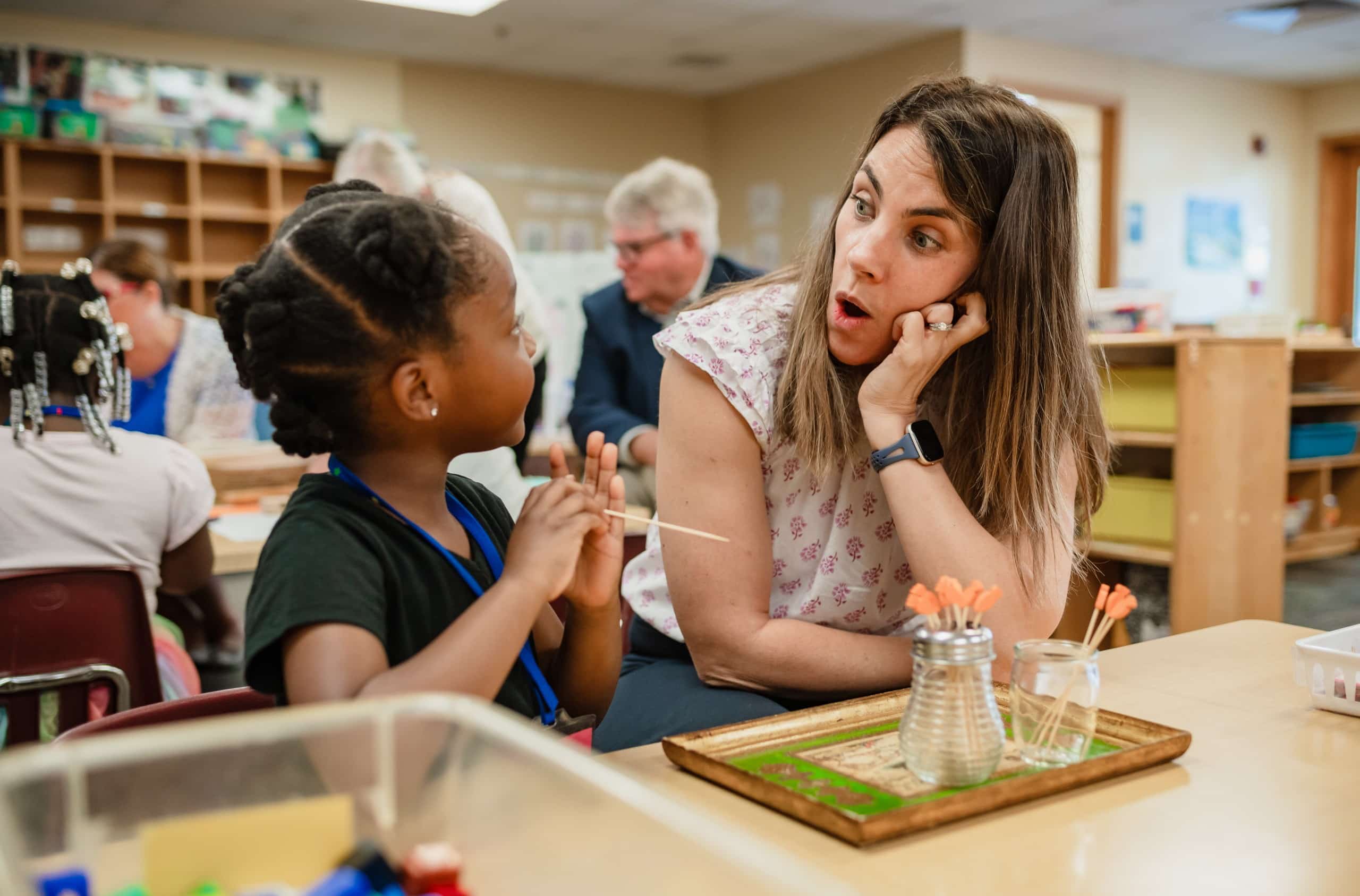 Inside a classroom for young students, a woman with long brown hair makes a surprised expression as she interacts with a young student, who is moving sticks from one to to another to learn counting.