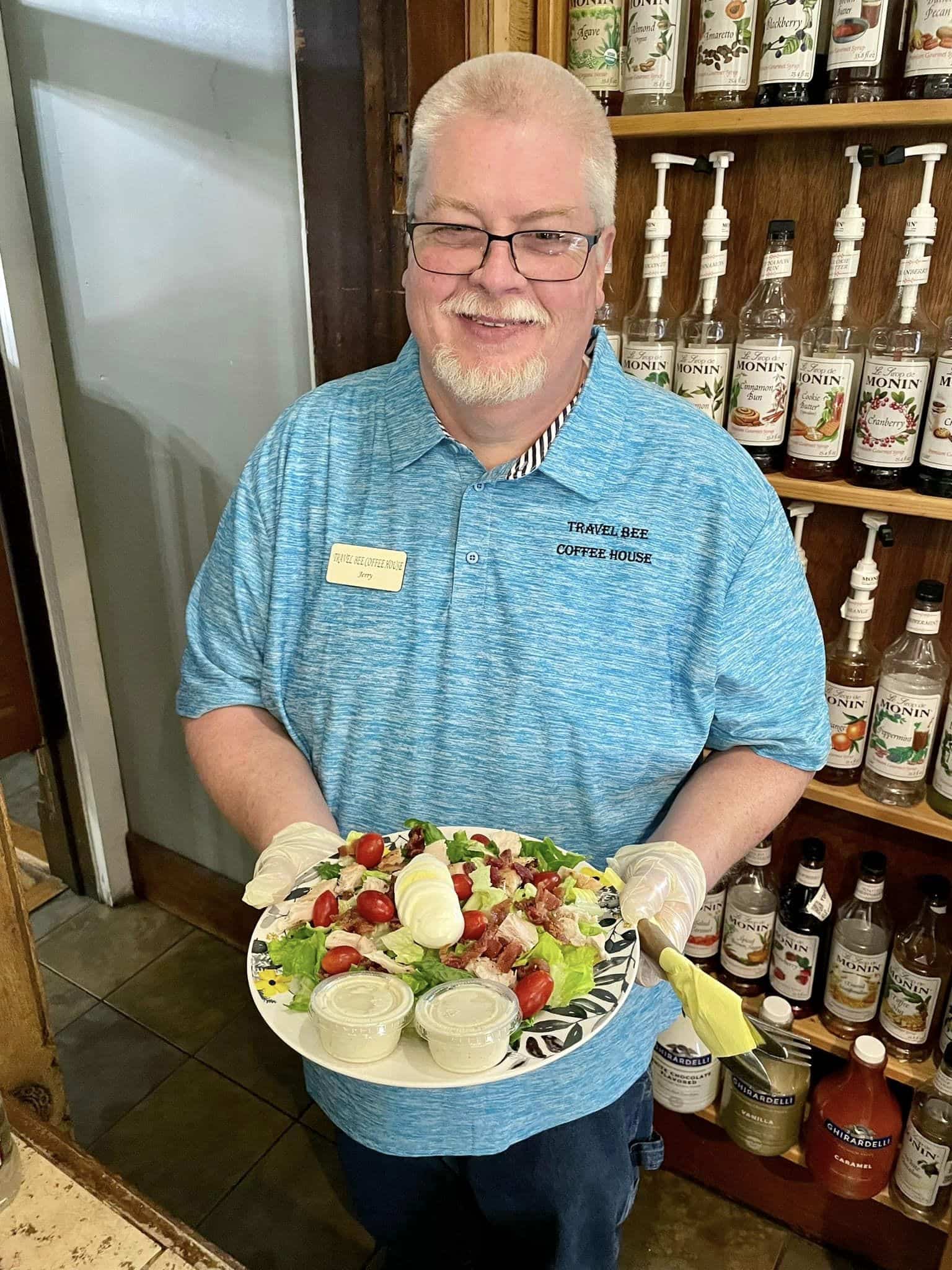 A man with white hair and facial hair in a light blue polo holds a white plate that features a salad.