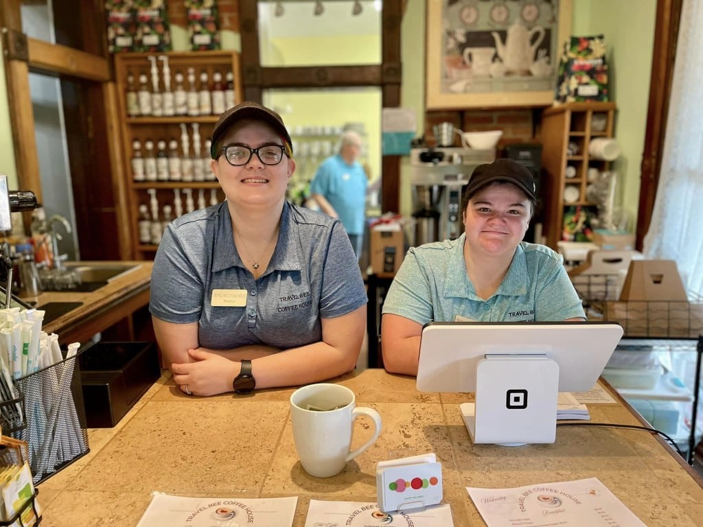 Inside a cozy coffee shop, two employees stand behind the front counter and register, smiling at the camera.