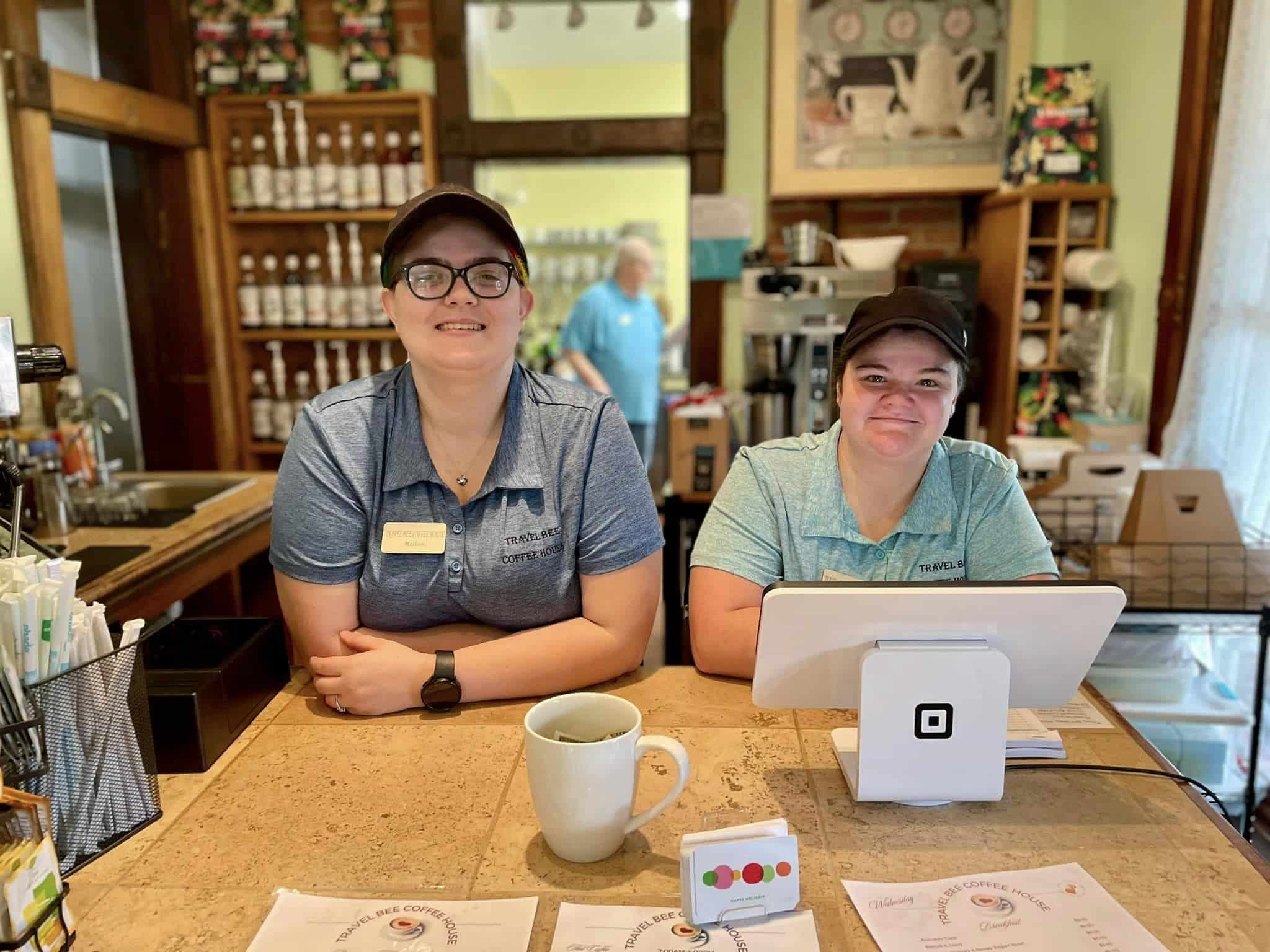 Inside a cozy coffee shop, two employees stand behind the front counter and register, smiling at the camera.