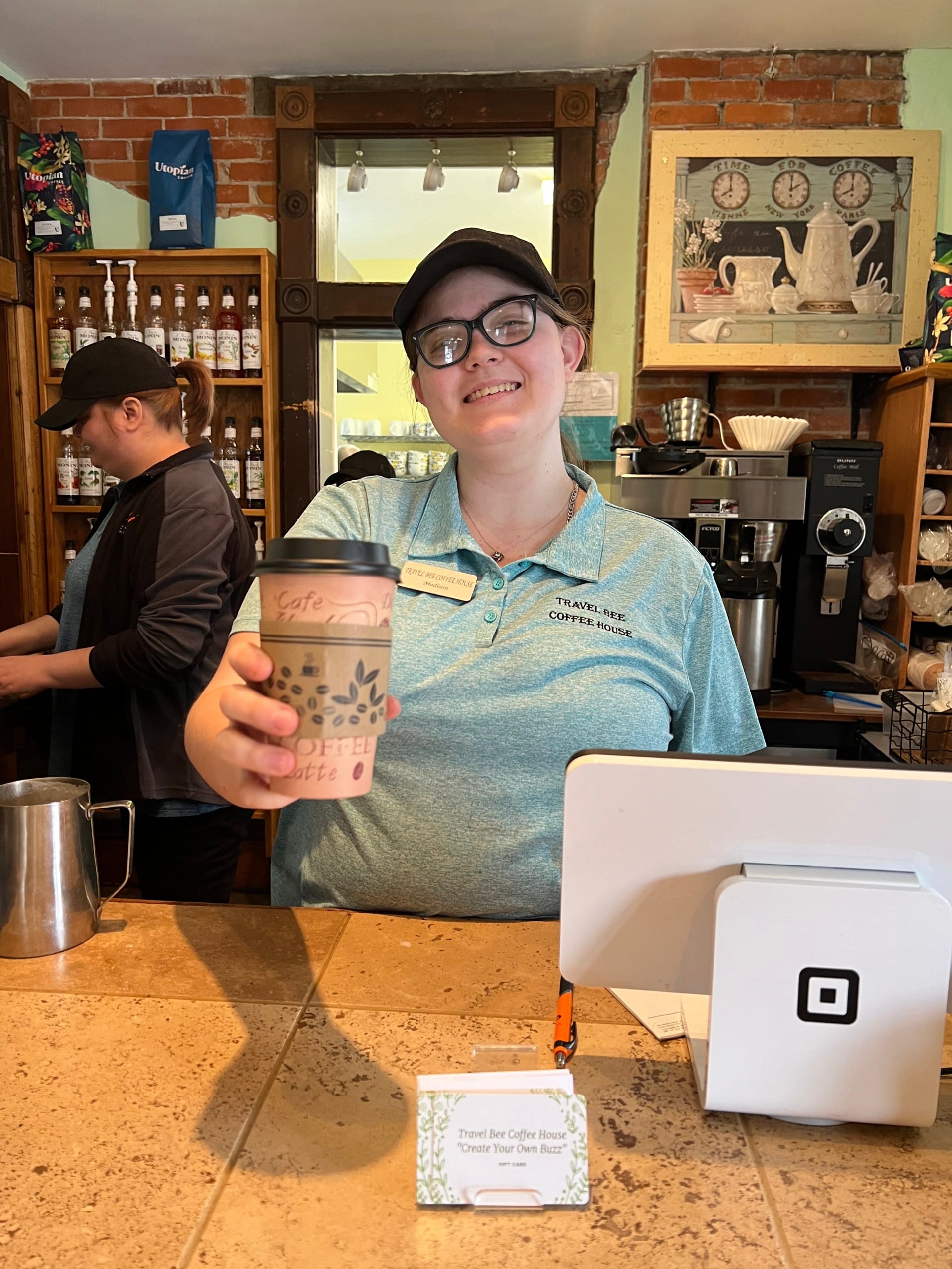 A woman wearing a black baseball hat and teal polo shirt, extends her arm out, holding a brown to-go coffee cup.