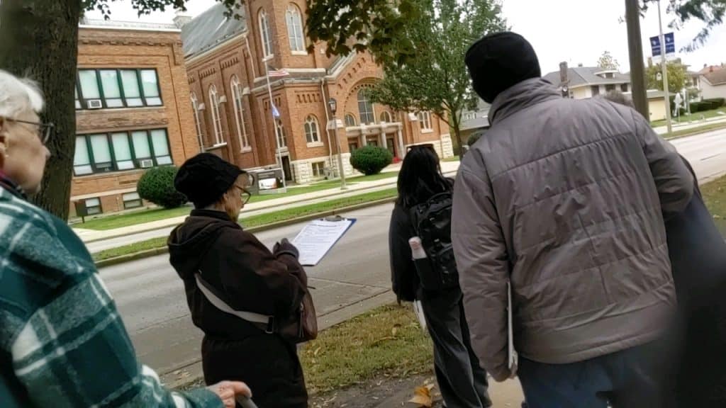 A group of people, one with a clip board, examine an uneven section of sidewalk in front of them.